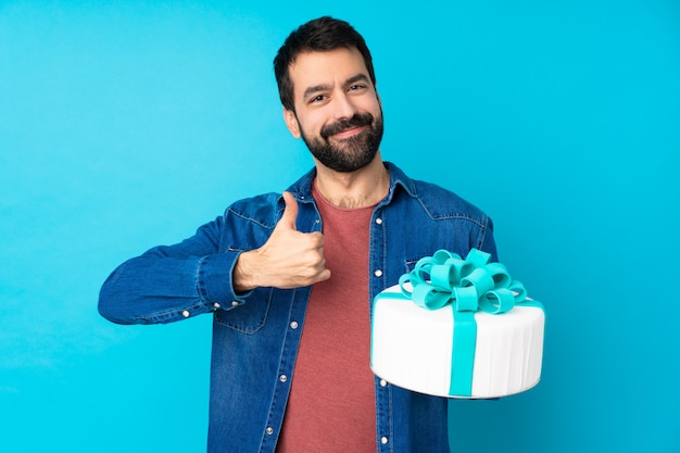 Young handsome man with a big cake over isolated blue wall giving a thumbs up gesture