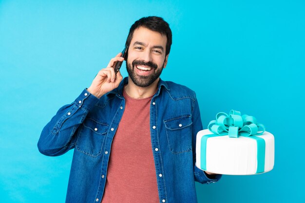 Young handsome man with a big cake over isolated blue wall keeping a conversation with the mobile phone with someone