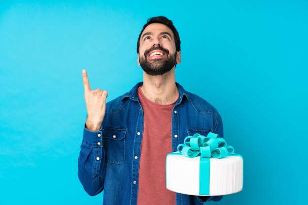 Giovane uomo bello con una grande torta sopra la parete blu isolata che indica su e sorpreso