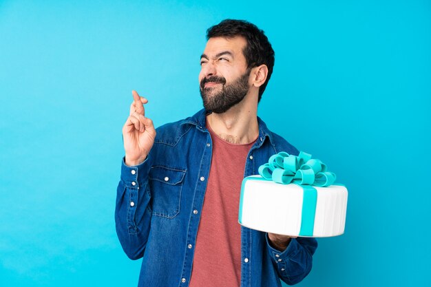 Young handsome man with a big cake over isolated blue wall with fingers crossing and wishing the best