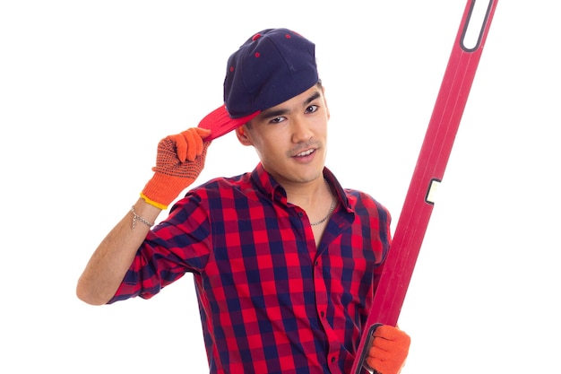 Young handsome man with black hair in red plaid shirt with orange gloves and blue snapback holding red level on white background in studio