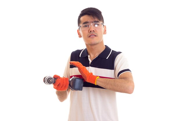 Young handsome man with black hair in white and blue tshirt with orange gloves and protective glasses holding grey electric screwdriver on white background in studio