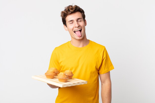 Young handsome man with cheerful and rebellious attitude, joking and sticking tongue out holding a muffins tray