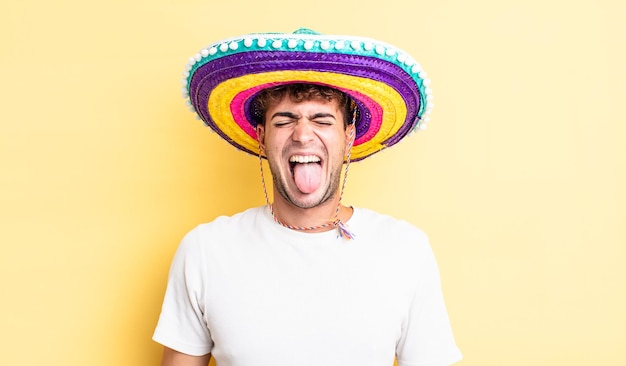 Photo young handsome man with cheerful and rebellious attitude, joking and sticking tongue out. mexican hat concept