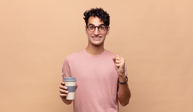 Young handsome man with a coffee feeling shocked, excited and happy, laughing and celebrating success, saying wow!