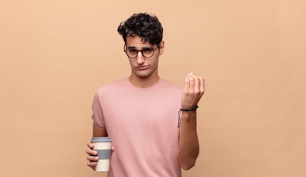 Young handsome man with a coffee making capice or money gesture, telling you to pay your debts!