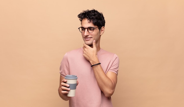 Young handsome man with a coffee smiling with a happy, confident expression with hand on chin, wondering and looking to the side