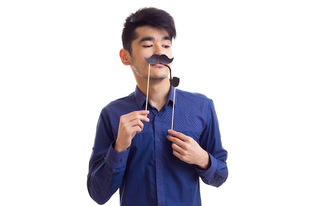 Young handsome man with dark hair in blue shirt holding cardboard stick of moustache in studio