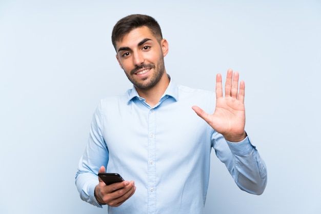 Giovane uomo bello con il suo cellulare che saluta con la mano con l'espressione felice