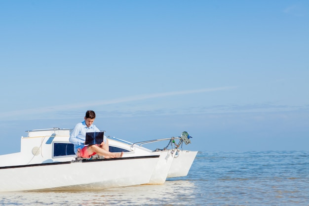 Uomo giovane e bello con il computer portatile sulla barca a vela. concetto di lavoro freelance