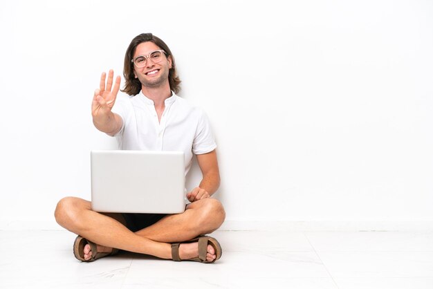 Young handsome man with a laptop sitting on the floor isolated on white background happy and counting three with fingers