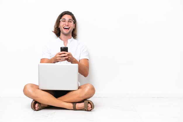Foto giovane uomo bello con un computer portatile seduto sul pavimento isolato su sfondo bianco sorpreso e inviando un messaggio