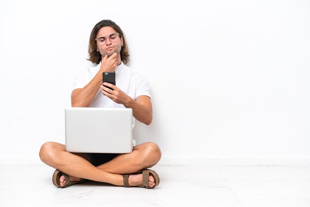 Foto giovane uomo bello con un computer portatile seduto sul pavimento isolato su sfondo bianco pensando e inviando un messaggio