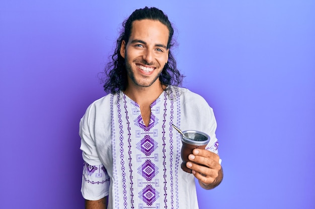 Young handsome man with long hair drinking mate infusion looking positive and happy standing and smiling with a confident smile showing teeth