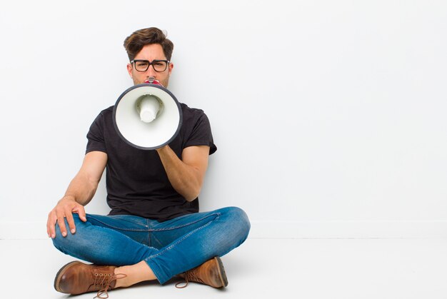 Young handsome man with a megaphone sitting on the floor sitting on the floor in a white room