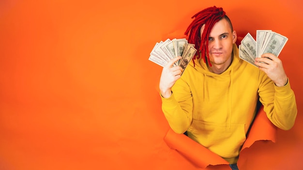 Young handsome man with money sticks out of hole of orange background Stylish hipster with dollar bills