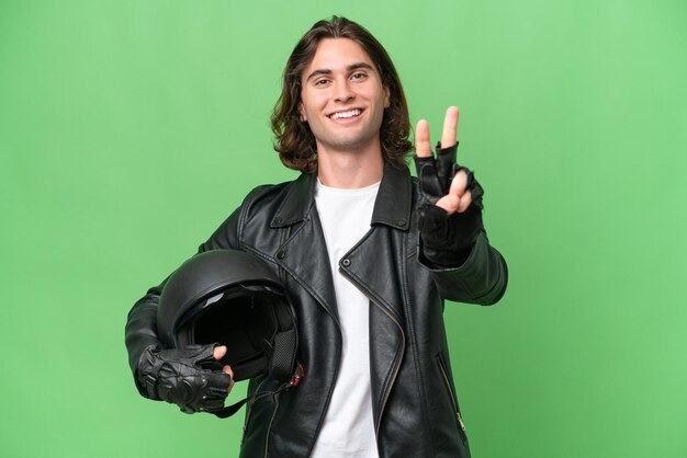 Young handsome man with a motorcycle helmet isolated on green chroma background smiling and showing victory sign