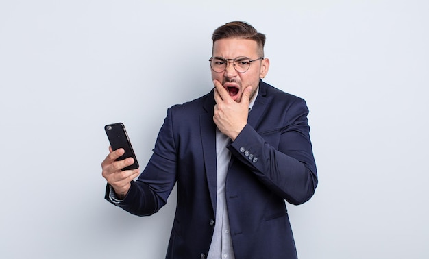 Young handsome man with mouth and eyes wide open and hand on chin business and smartphone concept