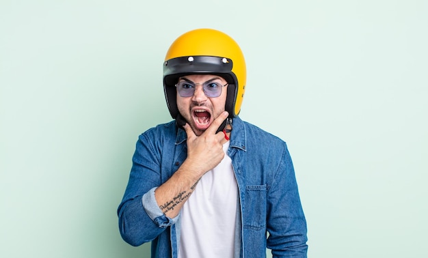 Giovane bell'uomo con la bocca e gli occhi spalancati e la mano sul mento. concetto di casco da moto