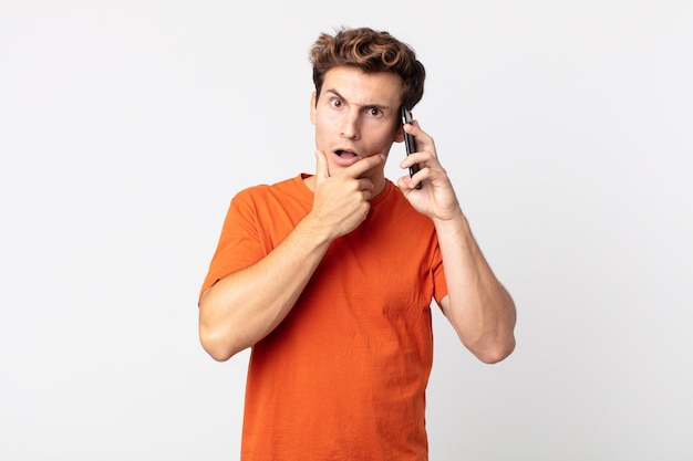 Young handsome man with mouth and eyes wide open and hand on chin and talking with a smart phone