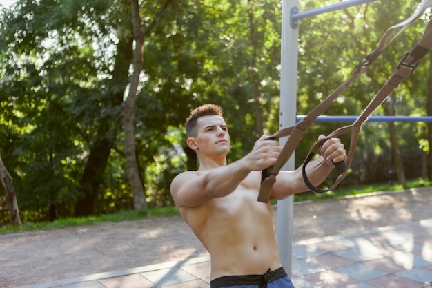 Young handsome man with naked torso trains with trx resistance system on outdoor sports ground. Healthy lifestyle concept