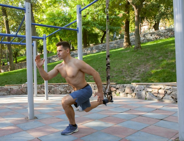 Young handsome man with naked torso trains with trx resistance system on outdoor sports ground. Healthy lifestyle concept