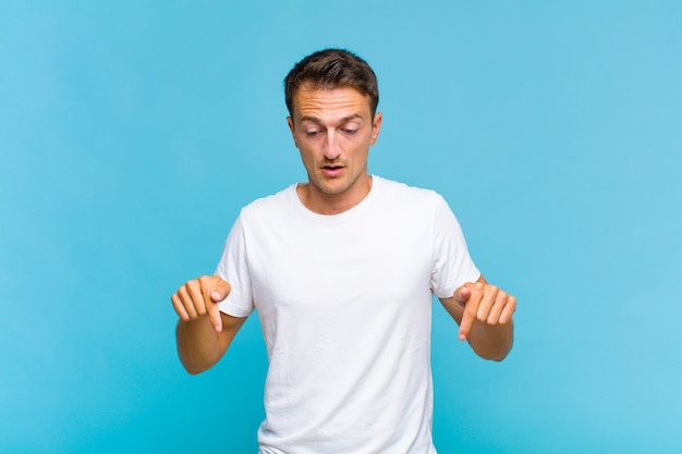 Young handsome man with open mouth pointing downwards with both hands, looking shocked, amazed and surprised