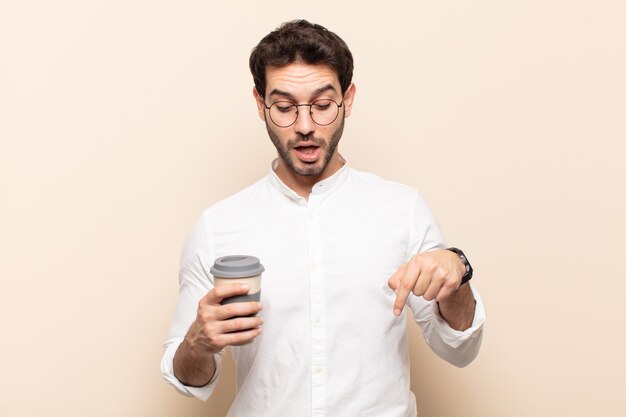 Young handsome man with open mouth pointing downwards with both hands, looking shocked, amazed and surprised