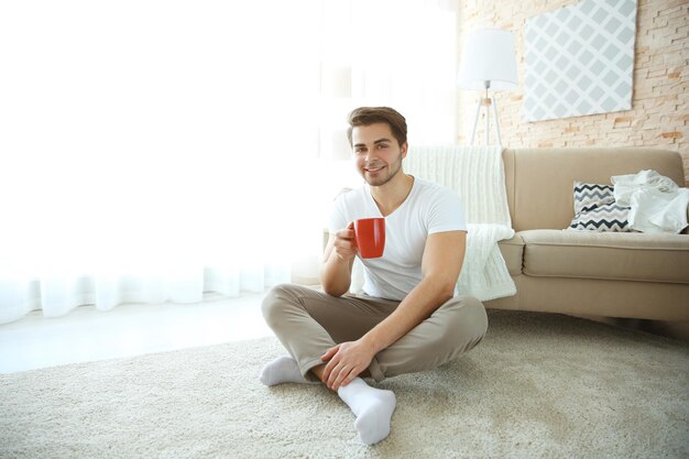 床に座ってコーヒーの赤いカップを持つ若いハンサムな男