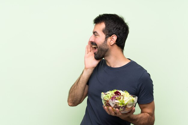 Young handsome man with salad over isolated green wall shouting with mouth wide open to the lateral