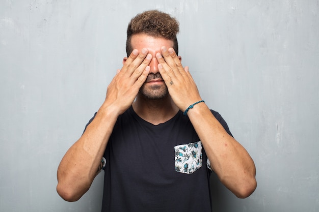 Photo young handsome man with a serious, scared, frightened expression, covering eyes with both hands.