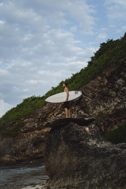 Foto giovane uomo bello con una tavola da surf su una roccia vicino all'oceano.