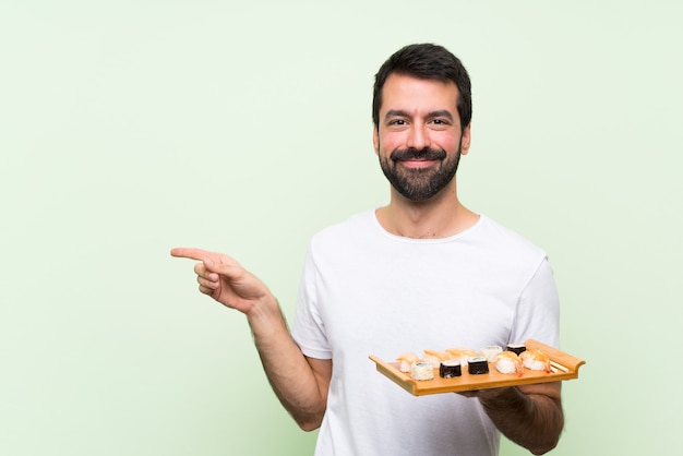 Young handsome man with sushi over isolated green wall pointing finger to the side