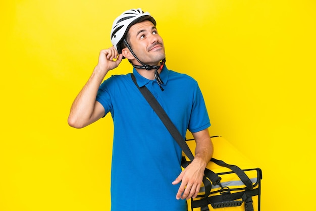 Young handsome man with thermal backpack over isolated yellow background having doubts and with confuse face expression