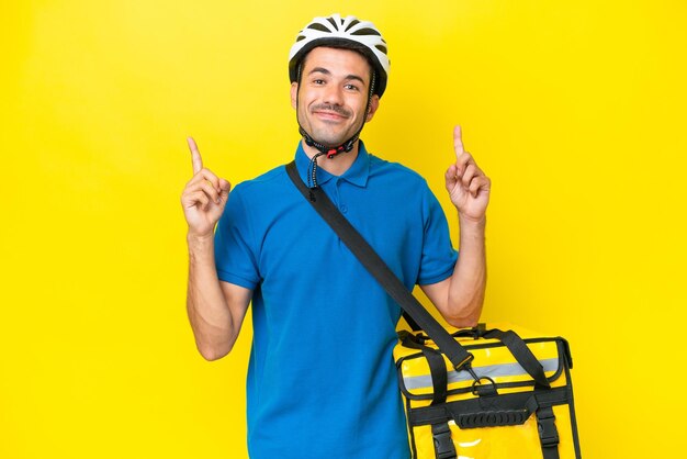 Young handsome man with thermal backpack over isolated yellow background pointing up a great idea