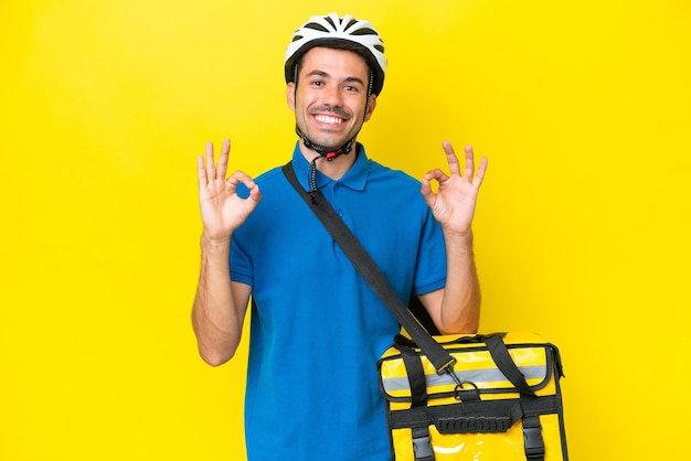 Молодой красивый мужчина с терморюкзаком на изолированном желтом фоне показывает пальцами знак "ок"