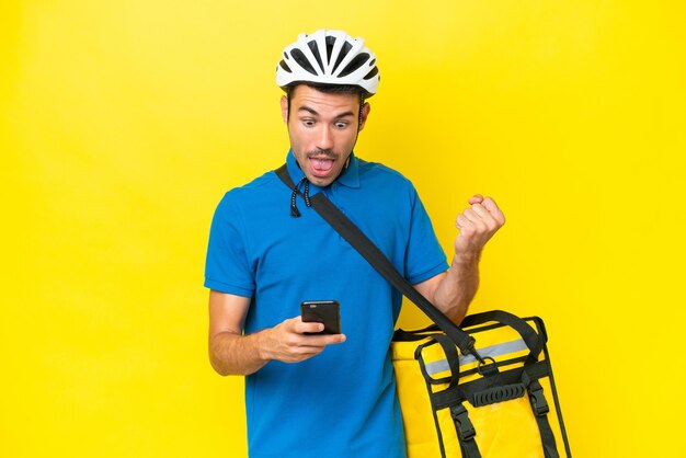 Young handsome man with thermal backpack over isolated yellow background surprised and sending a message