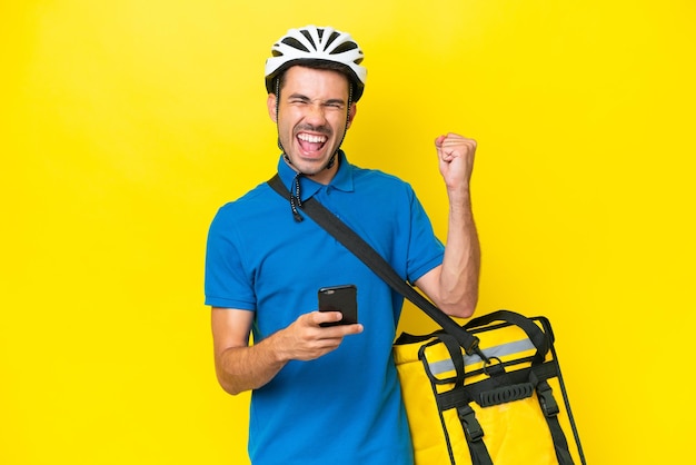 Young handsome man with thermal backpack over isolated yellow background with phone in victory position