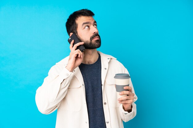 Young handsome man with white corduroy jacket over isolated blue wall holding coffee to take away and a mobile