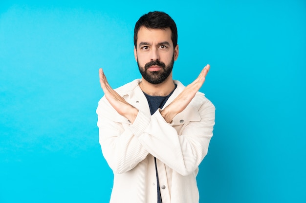 Young handsome man with white corduroy jacket over isolated blue wall making NO gesture