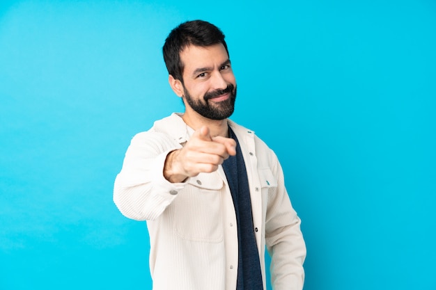 Young handsome man with white corduroy jacket over isolated blue wall points finger at you with a confident expression