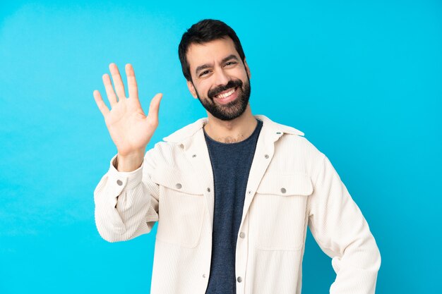 Young handsome man with white corduroy jacket over isolated blue wall saluting with hand with happy expression