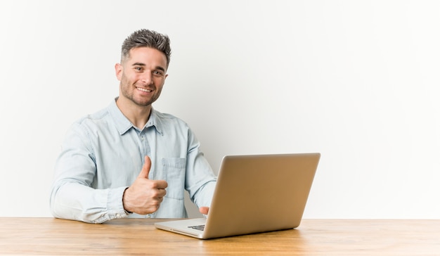 Giovane uomo bello che lavora con il suo computer portatile che sorride e che alza pollice in su