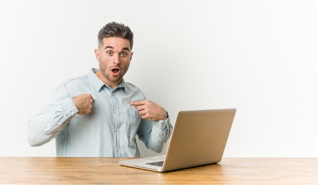 Young handsome man working with his laptop surprised pointing with finger, smiling broadly.