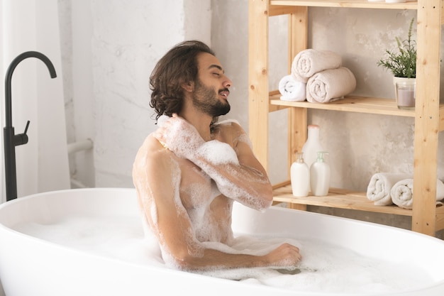 Giovane uomo nudo bello seduto nella vasca da bagno con acqua calda e schiuma, lavandosi il corpo e godersi la procedura