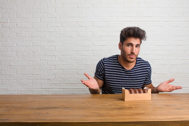 Young handsome and natural man sitting on a table doubting and shrugging shoulders