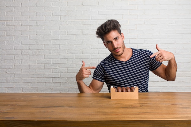 Young handsome and natural man sitting on a table proud and confident, pointing fingers, example to follow