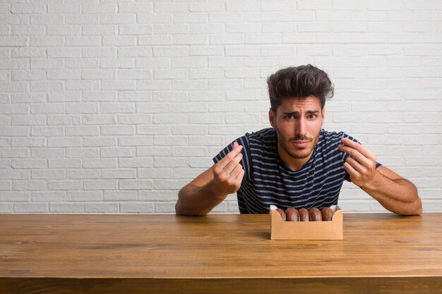 Giovane uomo bello e naturale seduto su un tavolo triste e depresso, facendo un gesto di necessità, restituendo alla carità, concetto di povertà e miseria. mangiare ciambelle al cioccolato.