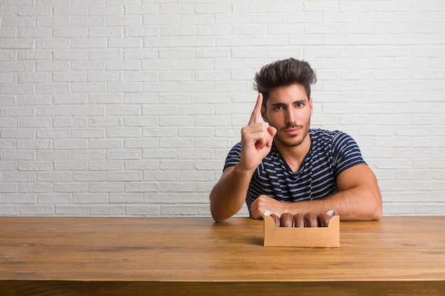 Young handsome and natural man sitting on a table showing number one, symbol of counting