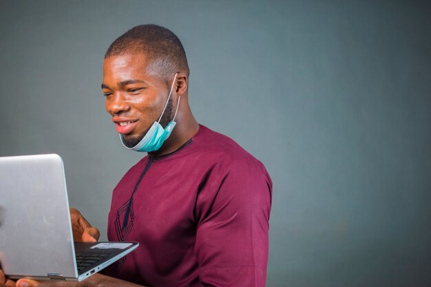 Young handsome nigerian man isolated over grey background wearing face mask and using his laptop because of virus outbreak.
